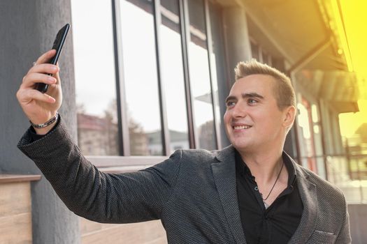 Businessman smiling man portrait of beautiful European appearance, in a jacket and shirt makes a selfie or talks on a video call on a smartphone on the street outdoor.