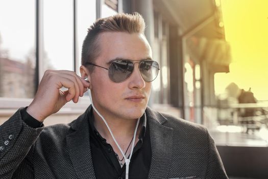 Portrait of a serious young guy of European appearance, an attractive businessman in sunglasses, listening to music in headphones on the street outdoor.