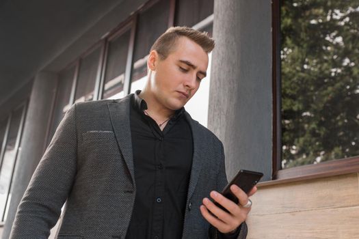A portrait of a stylish young guy of Caucasian appearance in a jacket and shirt uses a smartphone on the outdoor street. close-up.