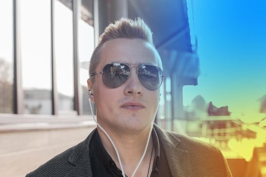 Portrait of a serious young guy of European appearance, an attractive businessman in sunglasses, listening to music in headphones on the street outdoor.