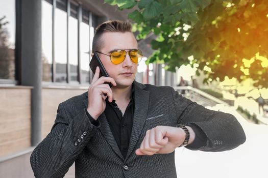 A young serious guy, businessman of European appearance portrait in sunglasses talks on a mobile phone or smartphone and looks at the time on a hand watch on the street outdoor.