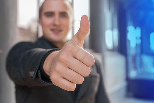 A young man shows a close-up of a thumbs up class against a street outdoor background.