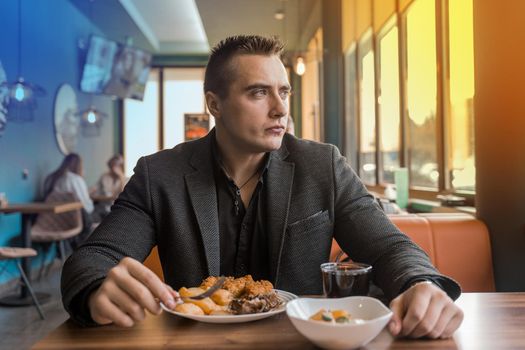 A serious, brooding stylish young man of European appearance, a businessman in a jacket sits at a table in a restaurant on a lunch break and looks out the window.