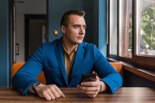 A brooding stylish, young businessman of Caucasian appearance portrait in a jacket and shirt sits at a table in a mobile phone and looks out the window.