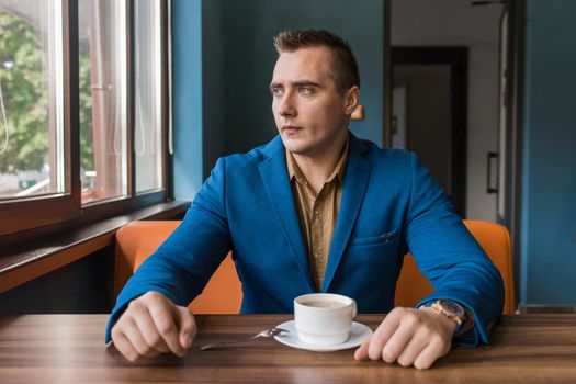 A young brooding, stylish businessman portrait of European appearance in a jacket and shirt sits at a table in a cafe for a cup of coffee on a lunch break and looks out the window.