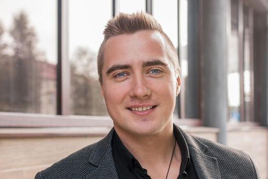 Close-up portrait of a positive smiling handsome guy of Caucasian appearance in a gray jacket and black shirt on the street outdoor.