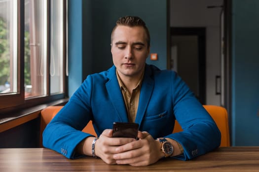 A young businessman of stylish European appearance in a blue suit and brown shirt sits at a table in a cafe in a mobile phone.