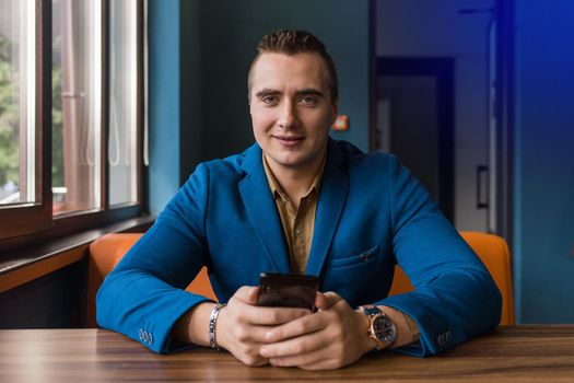 Stylish young, businessman of European appearance portrait of a man in a jacket and shirt sits at a table and uses a mobile phone in a cafe.