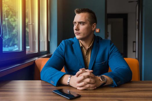 A brooding, stylish businessman portrait of European appearance in a jacket and shirt sits at a table in a cafe for a cup of coffee on a lunch break and looks out the window.