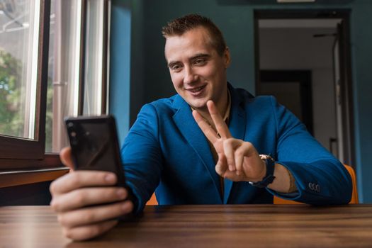 Smiling, positive businessman stylish young guy of Caucasian appearance portrait in jacket sits at a table in a cafe and fools around on a video link.