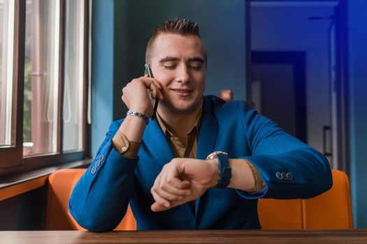 A cheerful, smiling man of stylish European appearance portrait of a businessman in a jacket talks on a mobile phone and looks at the time on the clock on his hand in a cafe.