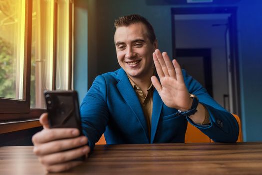 A young stylish guy, businessman of European appearance portrait sits at a table in a suit and talks via video communication in a smartphone transmitting greetings with a hand gesture.