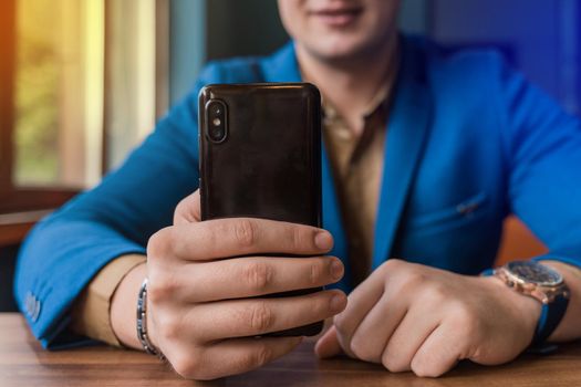 Close-up, businessman guy holds a black smartphone or mobile phone in his hand and takes a selfie or talks on a video link in a cafe.