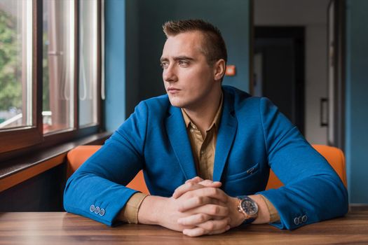 A brooding, young man of European appearance portrait businessman in jacket and shirt sits idly by at a table in a cafe and looks out the window.