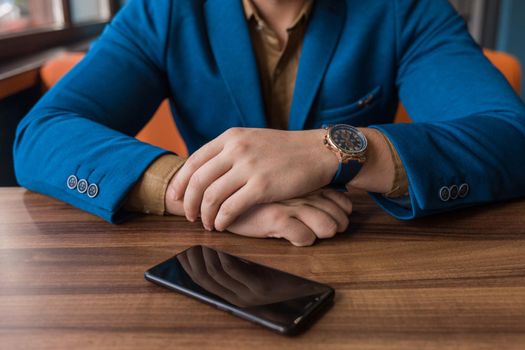 A businessman in a blue suit, jacket with a watch in his hands sits idly by next to a mobile phone or smartphone lying on a wooden table.