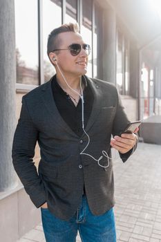 A young attractive smiling businessman of European appearance in a jacket and shirt style, wears sunglasses and listens to music in headphones from a smartphone on the street outdoor.