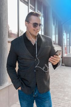 A young attractive businessman of European appearance in a jacket and shirt style, wears sunglasses and listens to music in headphones from a smartphone on the street outdoor.