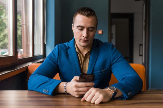 A young businessman of stylish European appearance in a blue suit and brown shirt sits at a table in a cafe in a mobile phone.