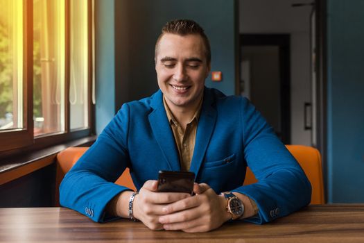 A smiling stylish, young businessman of Caucasian appearance portrait in a jacket and shirt sits at a table in a mobile phone and looks out the window.