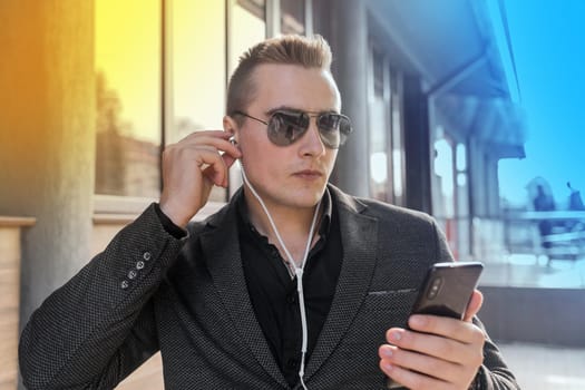 Portrait of a young stylish guy of Caucasian appearance of a businessman in sunglasses, jacket and shirt, adjusts his headphones with his hand listening to music from a mobile phone on the street outdoor.