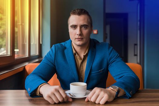 A collected adult, businessman portrait of Caucasian appearance in a blue jacket and brown shirt sits at a table in a cafe with a cup of coffee on a break.