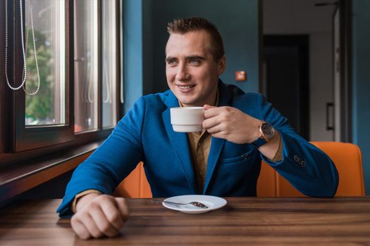 A stylish, positive businessman of Caucasian appearance, a man in a jacket and shirt sits at a table in a cafe on a coffee break and looks away or out the window.