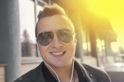 Portrait of a positive smiling young businessman of European appearance in sunglasses and headphones listening to music on the street outdoor.