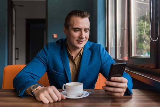 A young satisfied handsome guy businessman of European appearance portrait, uses a smartphone or mobile phone sitting in a cafe at a table on a coffee break.