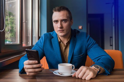 A seriously collected, brooding guy businessman of European appearance portrait sits at a table in a cafe on a coffee break and uses a smartphone or mobile phone.