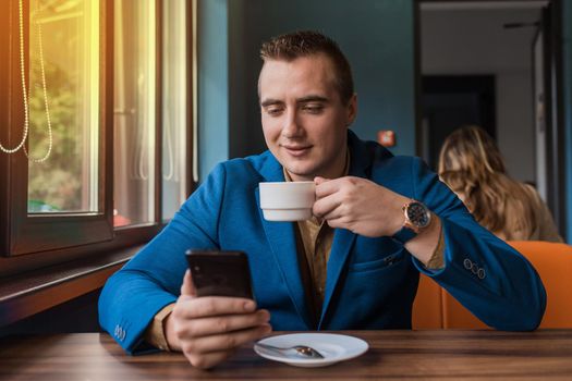 A satisfied, young businessman of Caucasian appearance cute portrait drinks coffee and uses a smartphone sitting at a table in a cafe on a lunch break.
