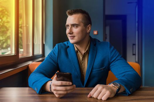 A positive stylish, young businessman of Caucasian appearance portrait in a jacket and shirt sits at a table in a mobile phone and looks out the window.