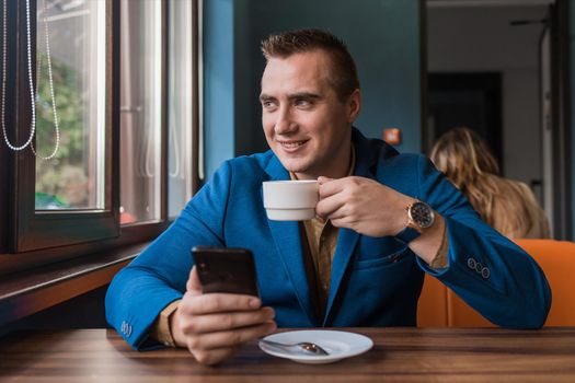 A young smiling positive handsome guy businessman of European appearance portrait, uses a smartphone or mobile phone sitting in a cafe at a table on a coffee break.