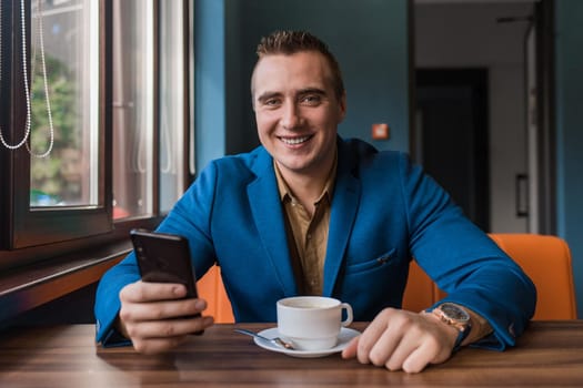 A young smiling positive handsome guy businessman of European appearance portrait, uses a smartphone or mobile phone sitting in a cafe at a table on a coffee break.