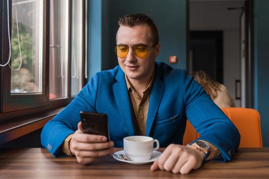 A stylish guy businessman of European appearance in sunglasses, a jacket and shirt sits at a table in a cafe on a coffee break, holds a smartphone or mobile phone in his hand.