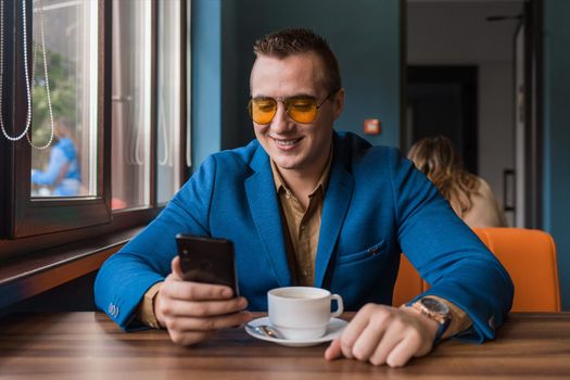 A stylish guy businessman of European appearance in sunglasses, a jacket and shirt sits at a table in a cafe on a coffee break, holds a smartphone or mobile phone in his hand.