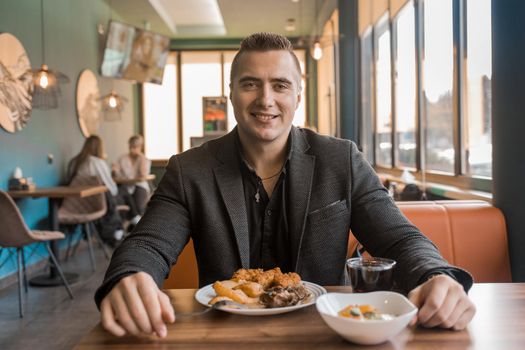 A young satisfied guy portrait of a businessman in a jacket sits at a table in a cafe on a lunch break.