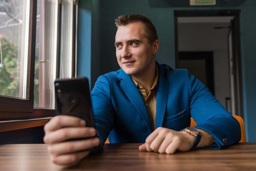 A positive, smiling young guy of European appearance, a stylish businessman, looks at a window and takes a selfie using mobile phone or smartphone.