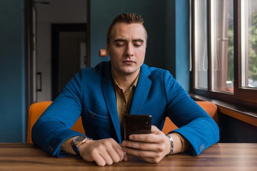 A young businessman, of stylish European appearance in a blue suit and brown shirt sits carried away at a table in a cafe in a mobile phone or smartphone.