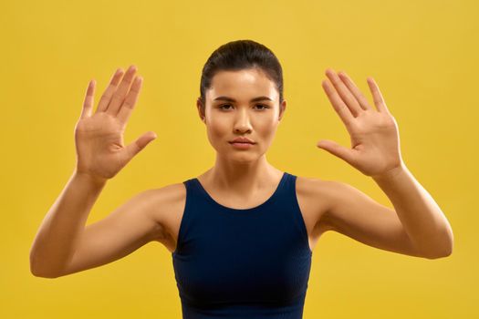 Front view of pretty girl raising hands, holding palms, looking at camera, squint. Slim brunette wearing sport suit, practicing yoga pose. Isolated on yellow studio background.