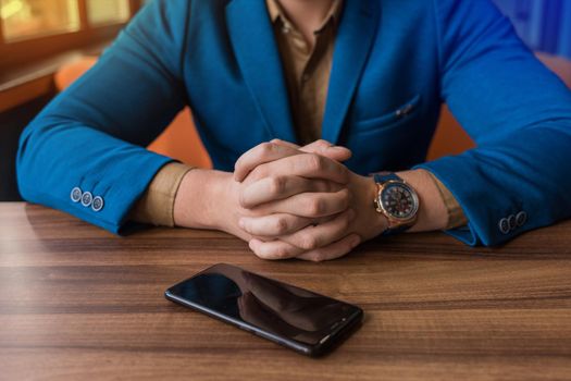 A businessman in a blue suit, jacket with a watch in his hands sits idly by next to a mobile phone or smartphone lying on a wooden table.