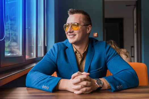 A satisfied happy guy of European appearance portrait of a stylish businessman in glasses, a suit, sits at a table in a cafe idly by and looks out the window.