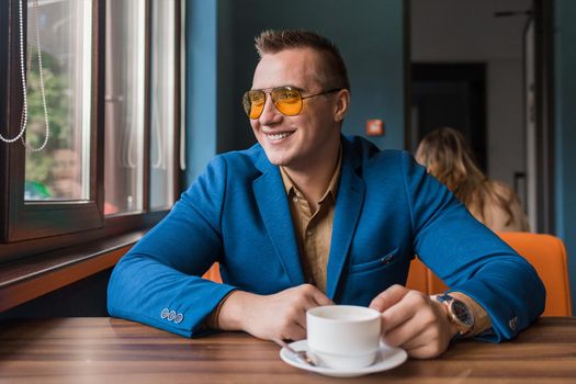 A positive smiling businessman a stylish portrait of Caucasian appearance in sunglasses, jacket and brown shirt, sits at a table on a coffee break and looks out the window in a cafe background.