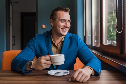 A stylish, positive businessman of Caucasian appearance, a man in a jacket and shirt sits at a table in a cafe on a coffee break and looks away or out the window.