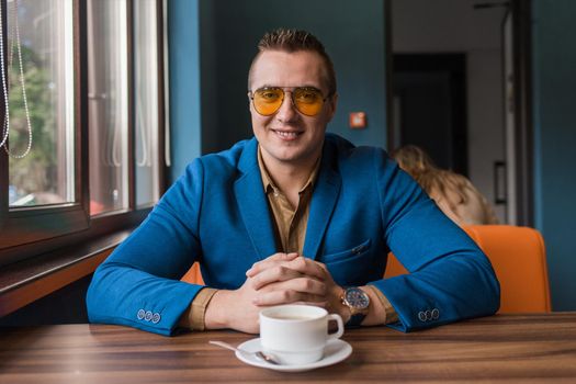 A positive smiling businessman a stylish portrait of Caucasian appearance in sunglasses, sits idly by at a table on a coffee break in a cafe background.