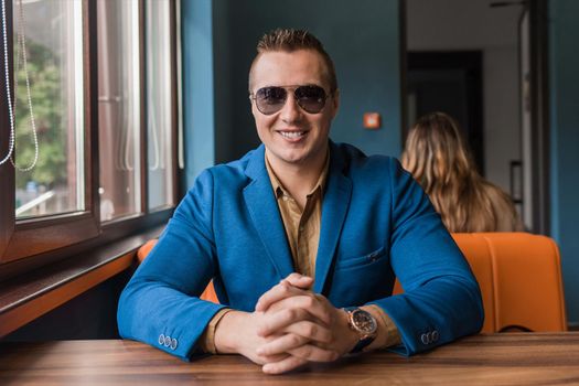 A young smiling businessman of European appearance stylish portrait with glasses sits at a table in a cafe idly by.