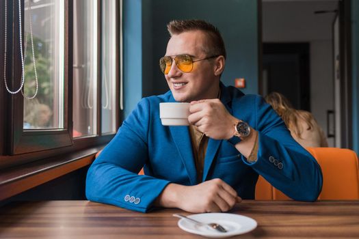 A positive smiling businessman a stylish portrait of Caucasian appearance in sunglasses, jacket and shirt, sits at a table on a coffee break and looks out the window in a cafe background.