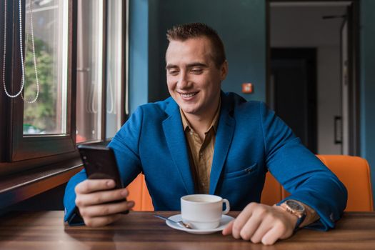 A young smiling positive handsome guy businessman of European appearance portrait, uses a smartphone or mobile phone sitting in a cafe at a table on a coffee break.