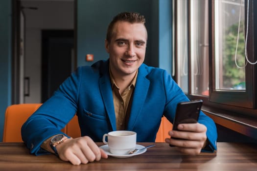 A young smiling positive handsome guy businessman of European appearance portrait, uses a smartphone or mobile phone sitting in a cafe at a table on a coffee break.