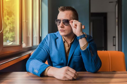 Business young businessman of Caucasian appearance stylish portrait adjusts sunglasses with his hands, looking out the window and sitting at table.