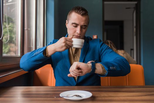 A busy business man a stylish businessman of Caucasian appearance in a suit and shirt watches the time and is late for negotiations, sitting at a cup of coffee at a table in a cafe.